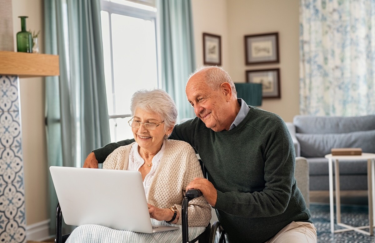 431495621 | © Rido - stock.adobe.com Lächelndes Seniorenpaar schaut zusammen auf einen Laptop, die Dame sitzt im Rollstuhl