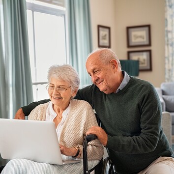 431495621 | © Rido - stock.adobe.com Lächelndes Seniorenpaar schaut zusammen auf einen Laptop, die Dame sitzt im Rollstuhl