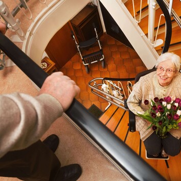 63423725 | © Ingo Bartussek - stock.adobe.com Eine ältere Dame fährt mit einem Treppenlift die Treppe hoch und hält dabei einen Blumenstrauß in der Hand