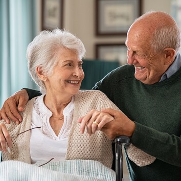 435778564 | © Rido - stock.adobe.com Seniorin im Rollstuhl und ein Senior daneben schauen und lächeln sich an