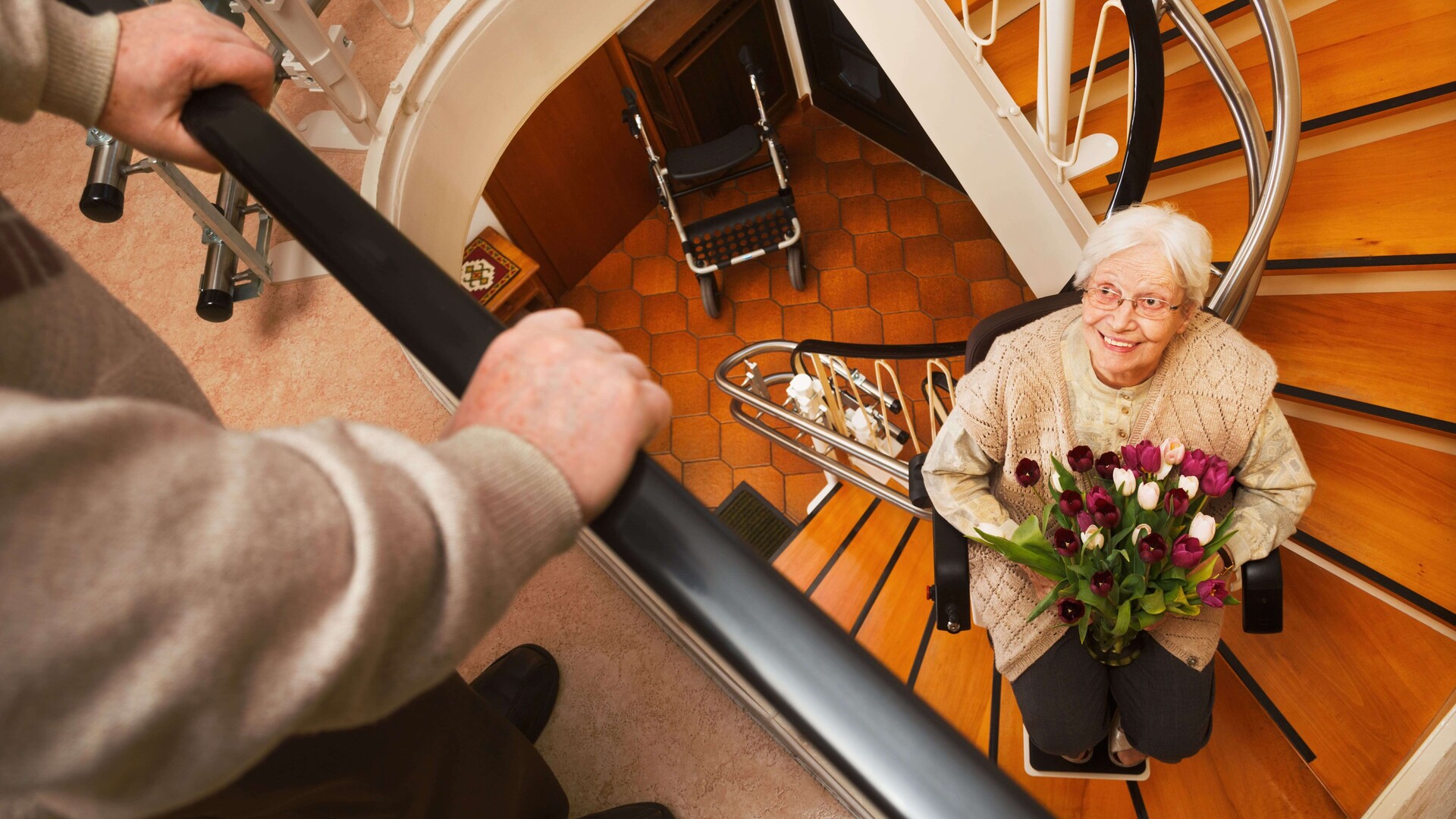 Eine ältere Dame fährt mit einem Treppenlift die Treppe hoch und hält dabei einen Blumenstrauß in der Hand