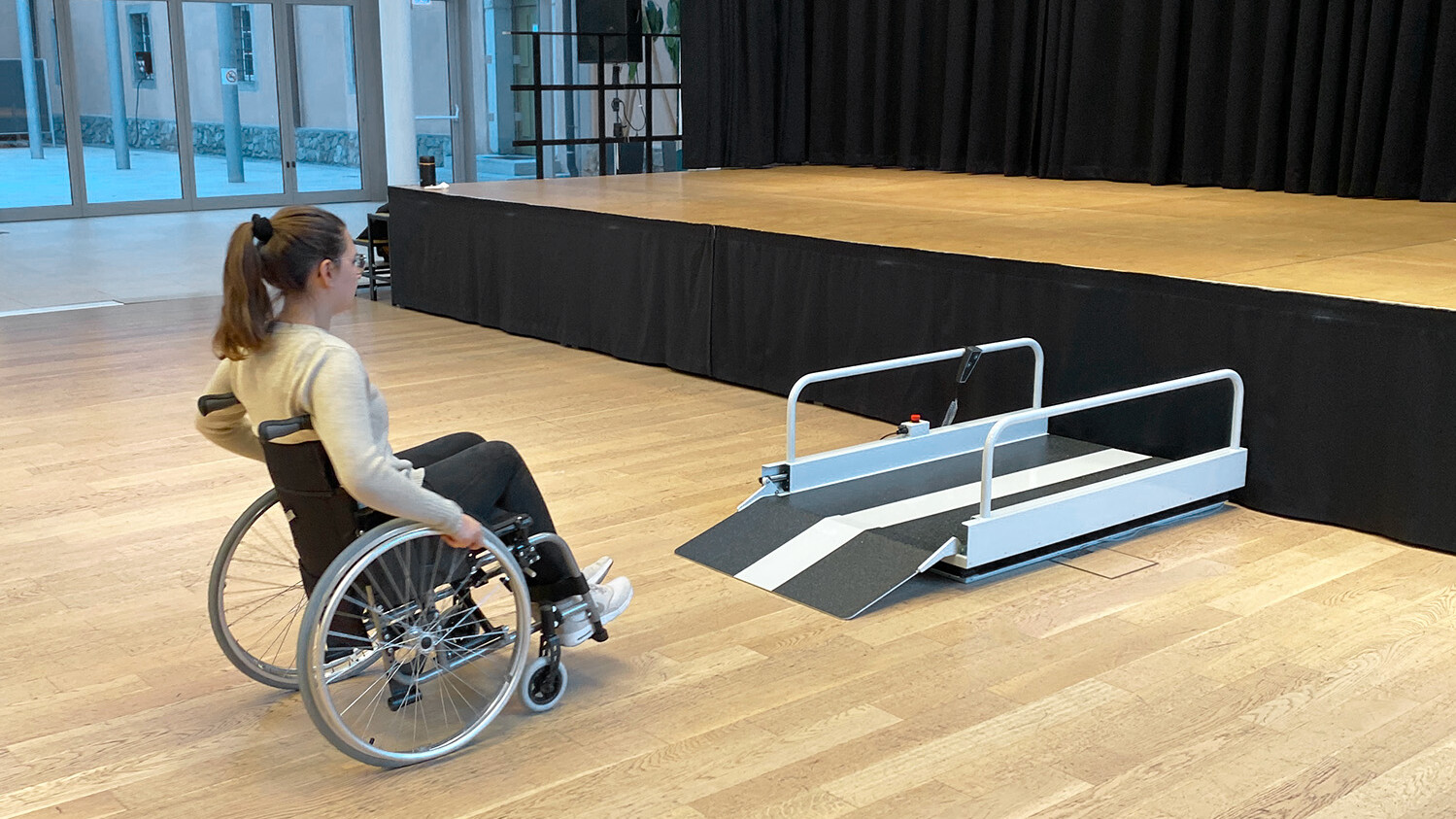 Eine Rollstuhlfahrerin nutzt den Lift Vertico Lite S, um auf eine Bühne zu gelangen