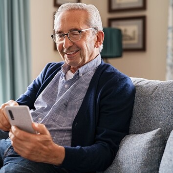 431495542 | © Rido - stock.adobe.com Senior lächelt, sitzt auf dem Sofa und schaut auf ein Smartphone