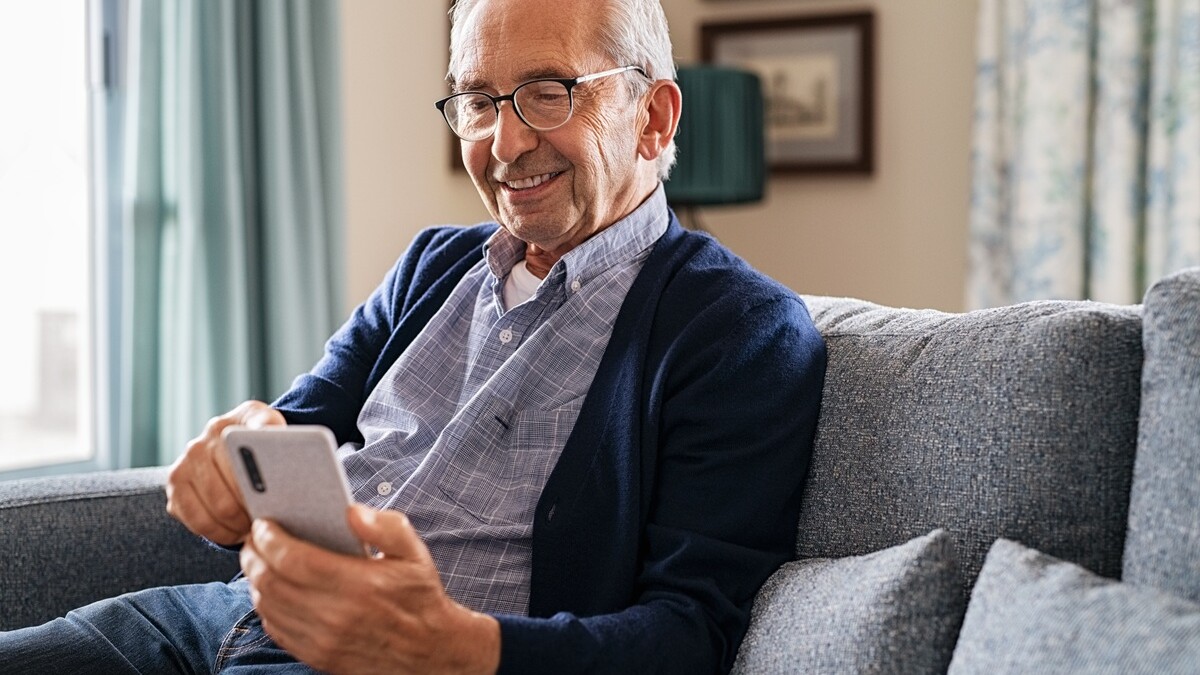Senior lächelt, sitzt auf dem Sofa und schaut auf ein Smartphone
